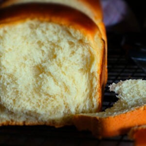 auf einem dunklen Gitter steht ein luftiges Brot. Dieses Brioche sieht fluffig weich aus. Eine Scheibe lehnt am Brot, die andere liegt rechts vor dem Brot