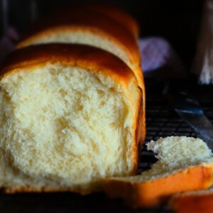 auf einem dunklen Gitter steht ein luftiges Brot. Dieses Brioche sieht fluffig weich aus. Eine Scheibe lehnt am Brot, die andere liegt rechts vor dem Brot