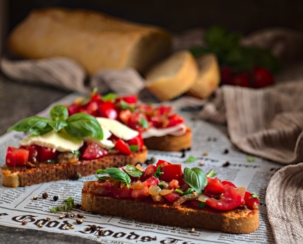 Bruchetta, Brote die auf einer Zeitung liegen, drei Stück belegt mit Tomaten, Käse und Basilikum. Im Hintergrund liegt ein Baguette auf einem Handtuch. das Gericht ist für den Optigrill