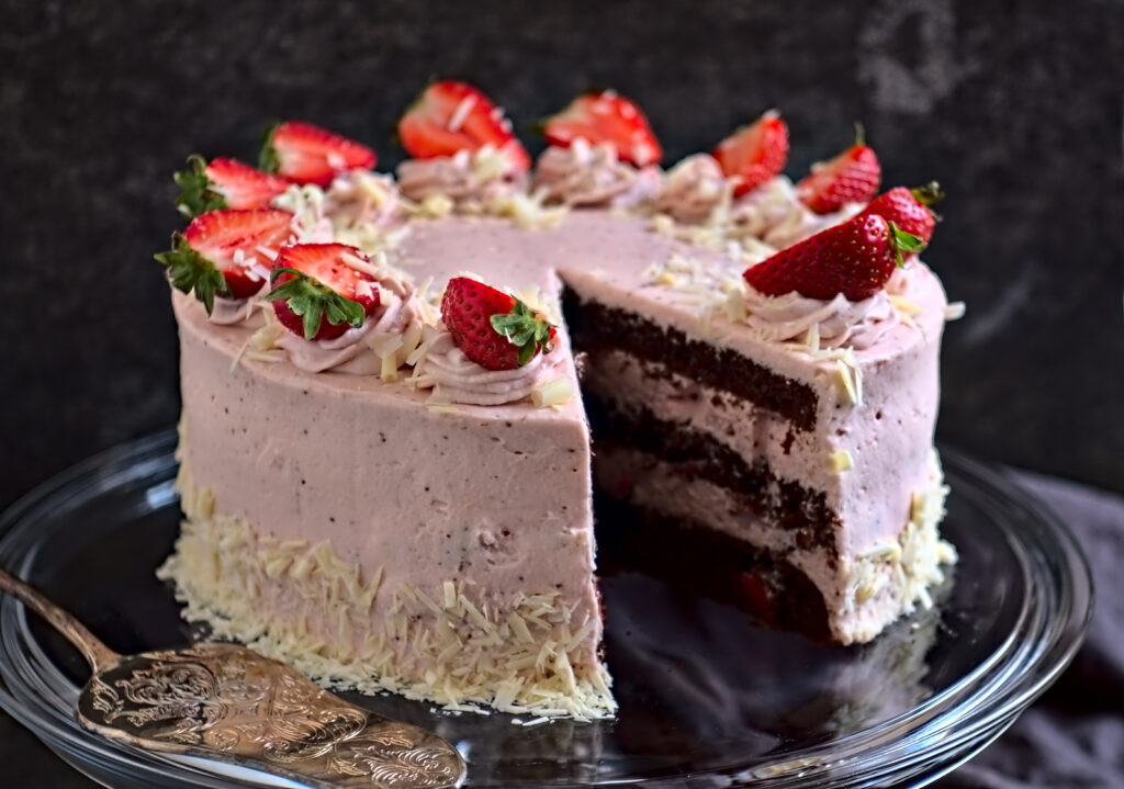 Eine cremige Erdbeer-Sahne-Torte wird auf einer Glasplatte präsentiert. Die angeschnittene Torte besteht aus drei Schichten Schokoboden und rosafarbener Erdbeercreme.