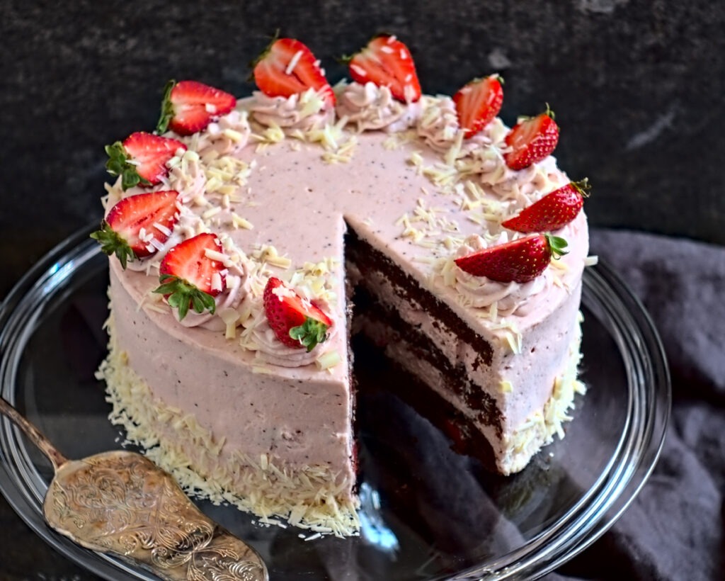 Eine Erdbeertorte aus drei Schichten Schokoladenkuchen und rosa Erdbeersahne steht auf einer Glasplatte. Die Erdbeer-Sahne-Torte ist verziert mit Sahnetupfen, weißen Schokoladenraspeln und aufgeschnittenen Erdbeeren.