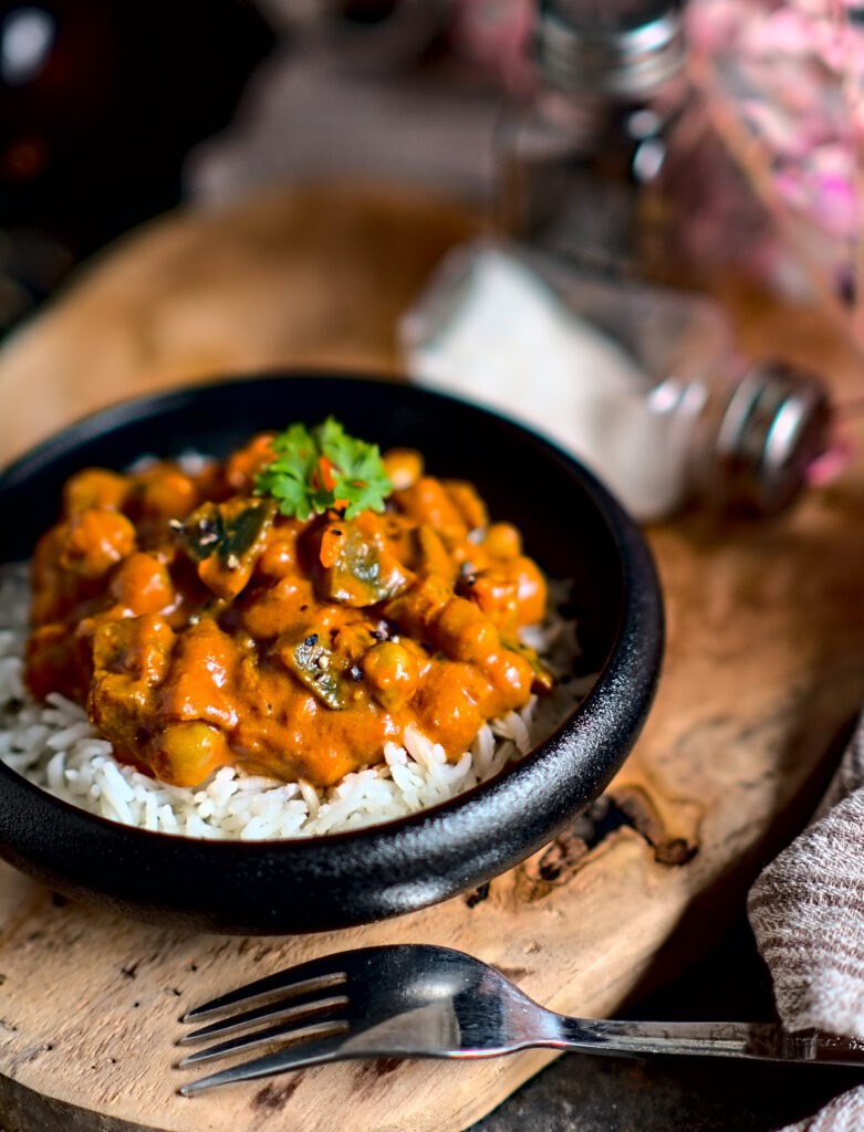 farbenfrohes Kichererbsen Curry mit Aubergine auf Reis in einer schwarzen Schüssel auf einem Holzbrett