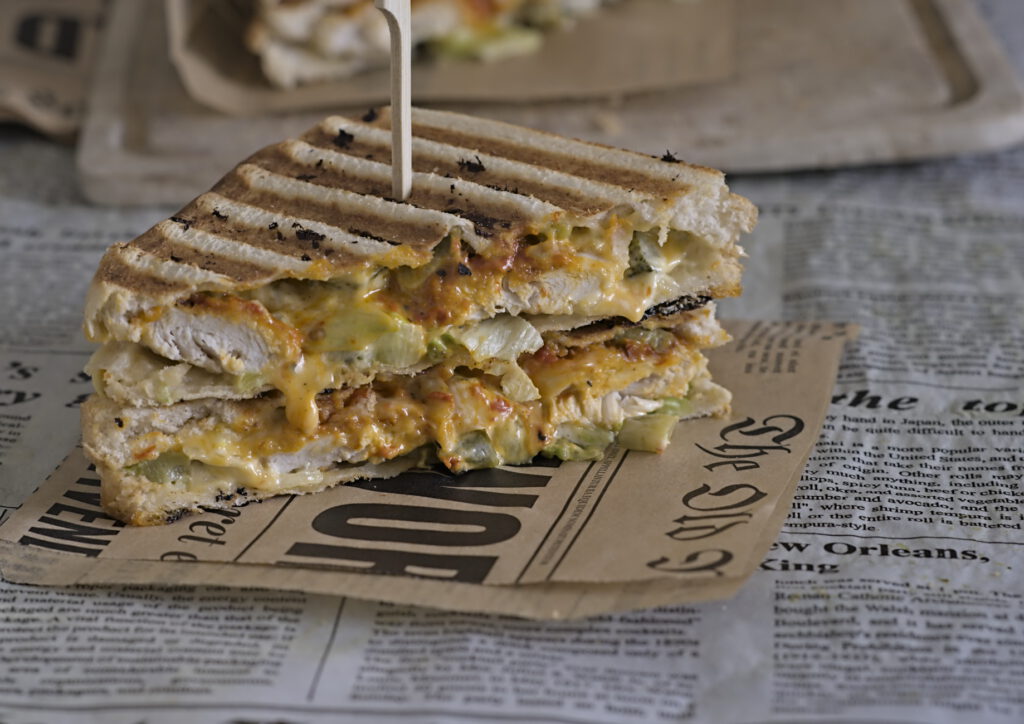 Ein Sandwich liegt auf einem Holzbrett. 1 Sandwich ist im Hintergrund zu sehen. Zu sehen ist der Salat, Chicken und eine cremige Soße