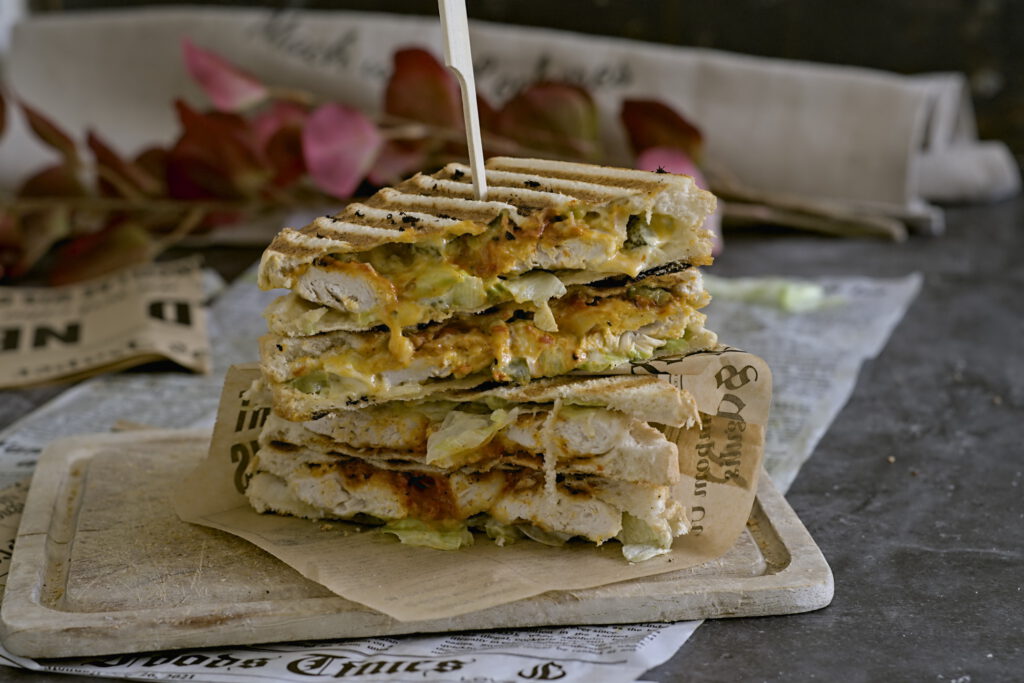 Ein Sandwich liegt auf einem Holzbrett. 2 Stück aufeinander gestapelt. Zu sehen ist der Salat, Chicken und eine cremige Soße