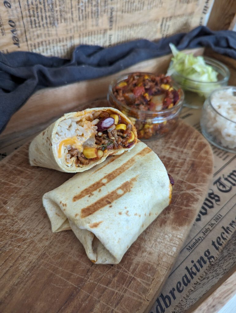 Auf einem Brett liegt ein Burrito einmal durchgeschnitten. Man sieht schön den Inhalt dieses Wraps. Daneben stehen einige Gefäße mit verschiedenen Zutaten.