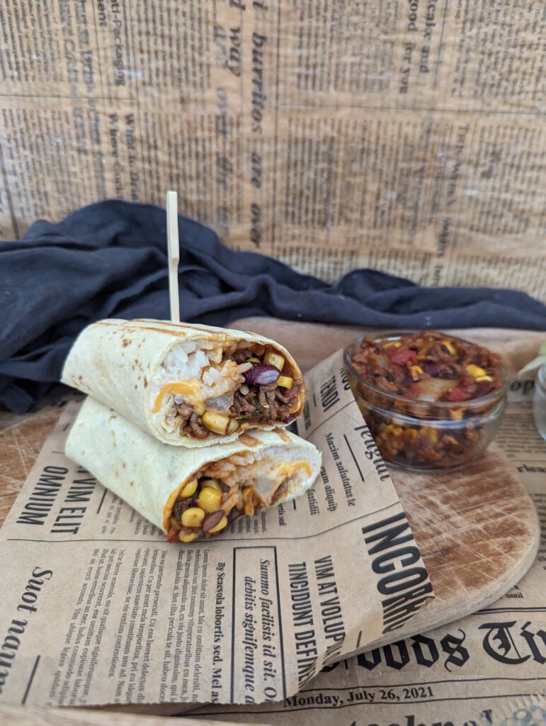 Auf einem Brett liegt ein Burrito einmal durchgeschnitten. Man sieht schön den Inhalt dieses Wraps. Daneben stehen einige Gefäße mit verschiedenen Zutaten.