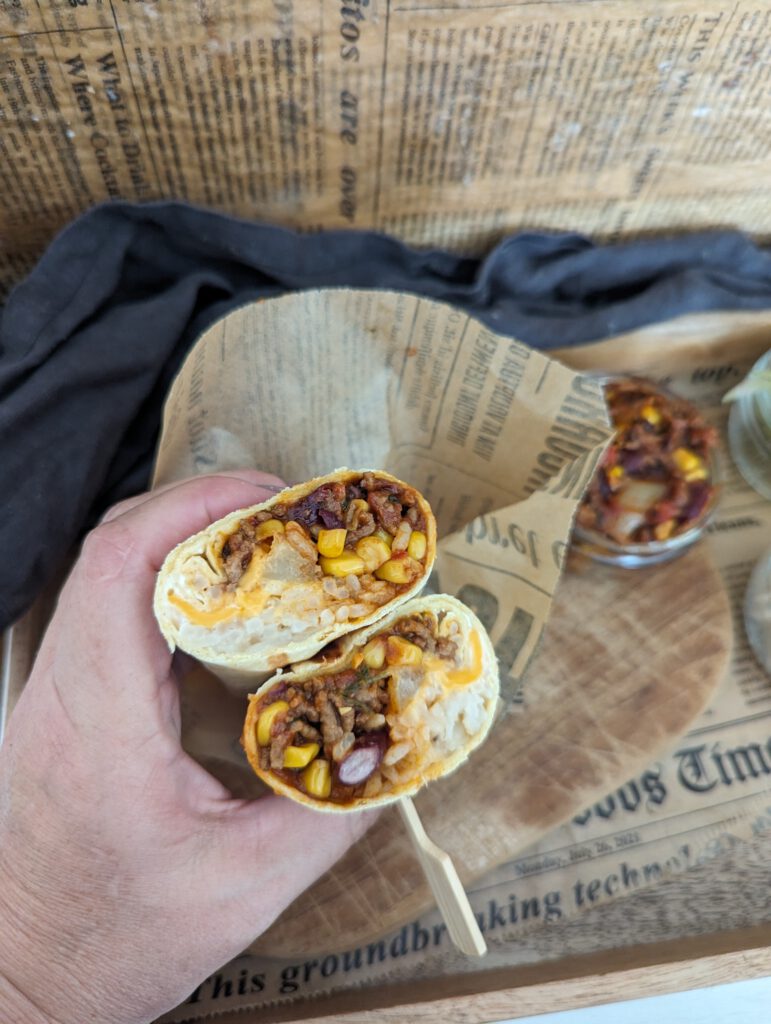 Auf einem Brett liegt ein Burrito einmal durchgeschnitten. Man sieht schön den Inhalt dieses Wraps. Der Wrap wurde in die Hand genommen