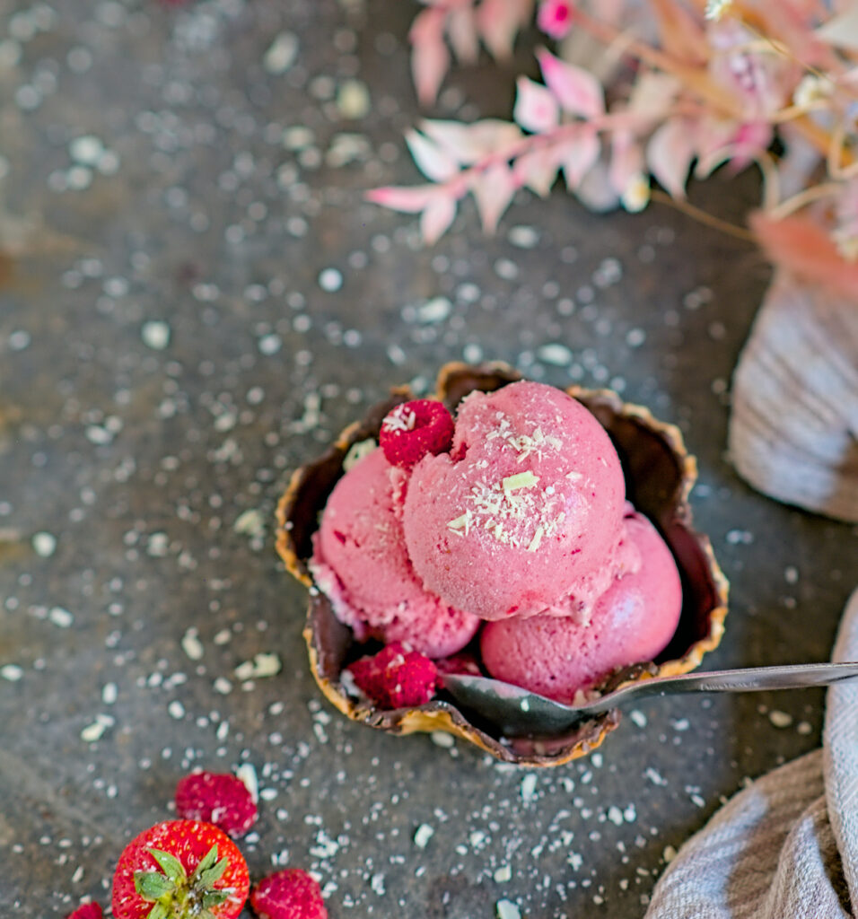 Drei Kugeln Fruchteis ohne Eismaschine in einem Waffelbecher mit Erdbeeren, Himbeeren und weißer Schokolade angerichtet