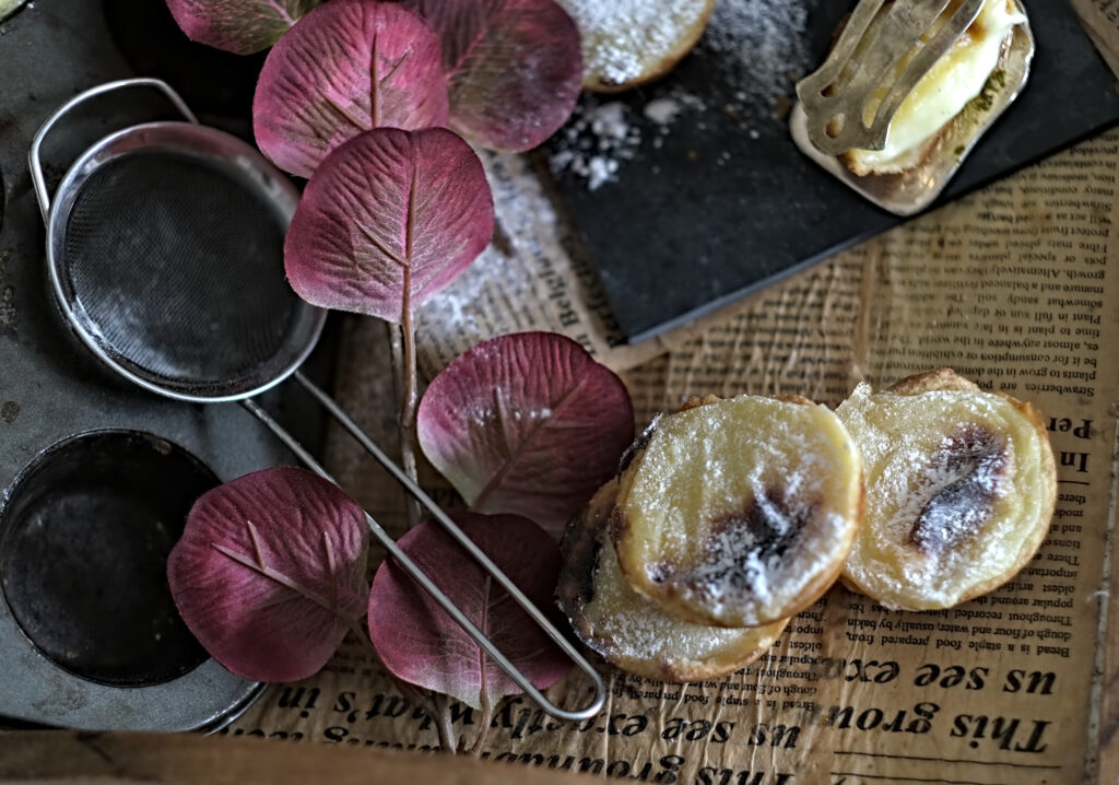 Pastel de Nata auf einer Zeitungsplatte, mit Blümchen und Sieb