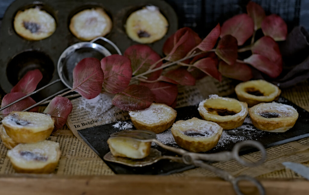 Pastel de Nata auf einem schwarzem Brett und in einer Muffinform. mit roten Blättern, Sieb und Gebäckstange