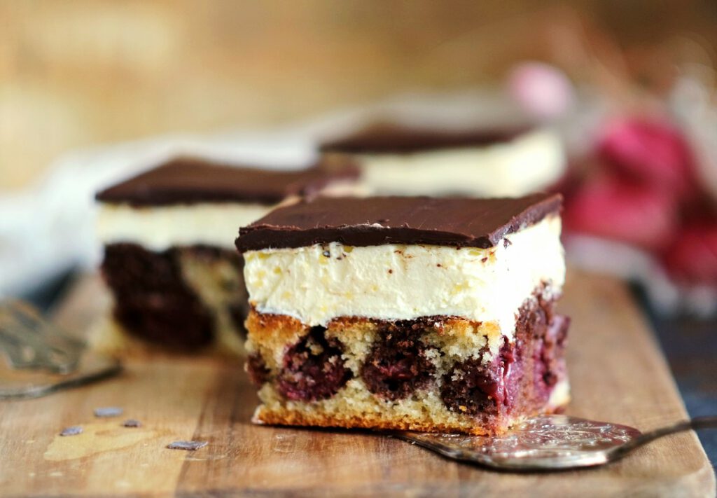 Nahaufnahme von einem angeschnittenen Stück Donauwelle. Die knackige Schokoglasur bricht leicht über der dicken, hellen Buttercreme-Schicht und dem marmorierten Kirschkuchen