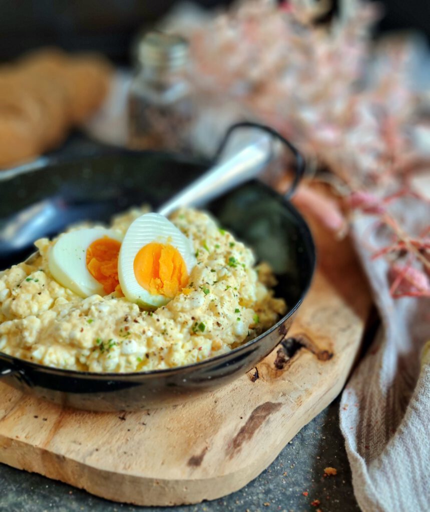 Das Bild zeigt den leichten Eiersalat in einer schwarzen Pfanne auf einem rustikalen Holzbrett, garniert mit frischen Kräutern und einem halbierten, hartgekochten Ei