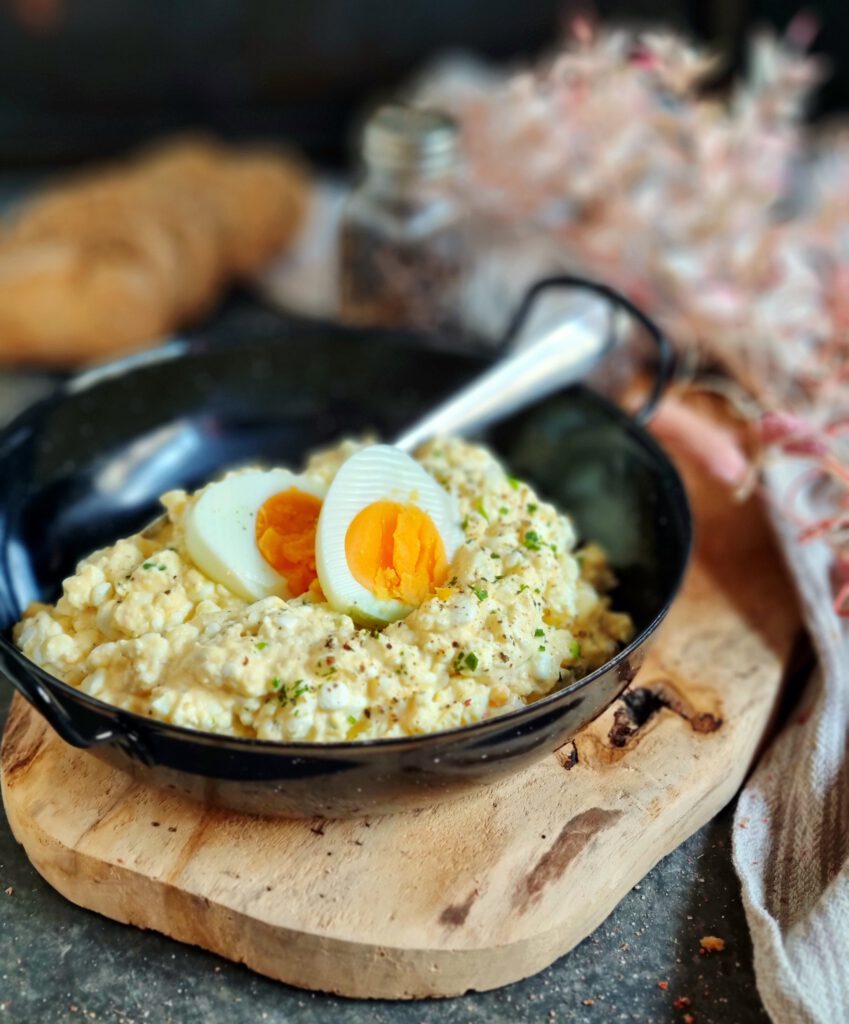 Gesunder Hüttenkäse-Eiersalat in einer schwarzen Gusseisenpfanne auf einem Holzbrett, getoppt mit einem halbierten Ei und Kräutern