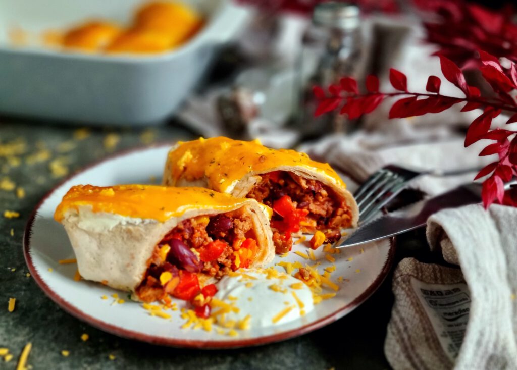 Mit Cheddar überbackene Enchiladas mit Hackfleischfüllung auf einem hellgrauen Teller mit einem Klecks Schmand, serviert auf einer Schieferplatte