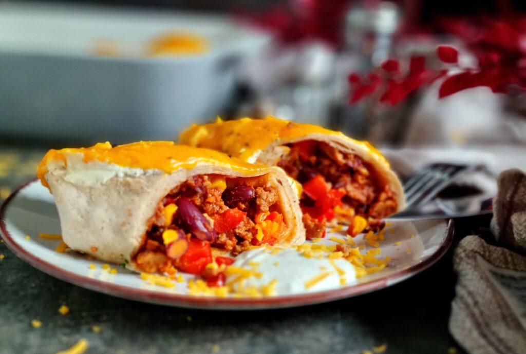 Nahaufnahme von goldbraun überbackenen Enchiladas, gefüllt mit einer saftigen Mischung aus Hackfleisch, Kidneybohnen, Mais und roter Paprika in Tomatensoße. Die Enchiladas sind mit geschmolzenem Cheddar überzogen und mit einem glatten Klecks Schmand garniert.