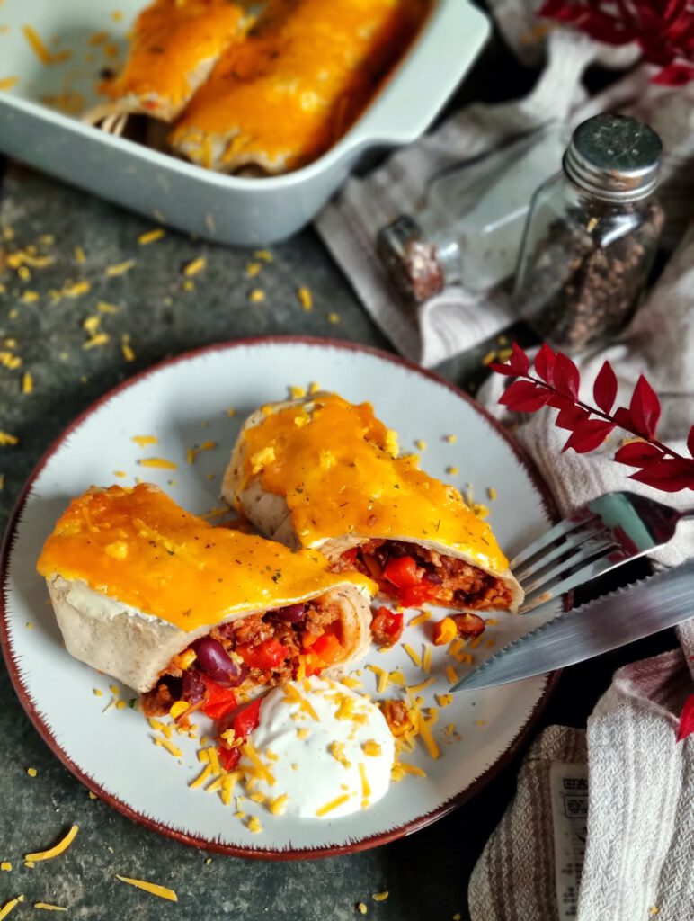 Portion überbackene Hackfleisch-Enchiladas mit geschmolzenem Cheddar und einem Klecks Schmand auf einem hellgrauen Teller